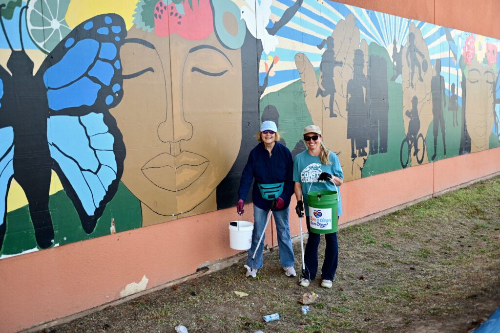 Two volunteers at I Love A Clean San Diego's Creek to Bay cleanup pick up trash in front of a mural.